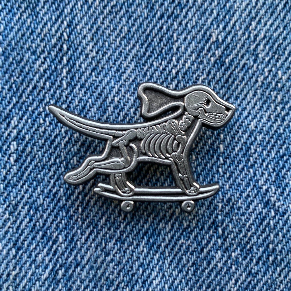 Silver pin depicting a running skeleton dog riding a skateboard